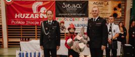 Prezes Zarządu Gminnego OSP oraz Komendant Powiatowy PSP wręczają nagrodę uczestnikowi zawodów. Zdjęcie wykonane w hali sportowej.