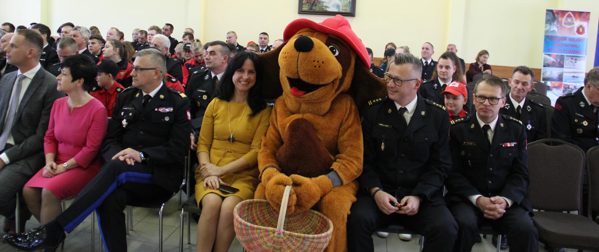 Na widowni. W pierwszym rzędzie goście i duża maskotka strażaka, która siedzi pomiędzy nimi. 