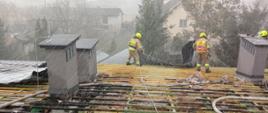 Na zdjęciu strażacy w hełmach i ubraniach specjalnych piaskowych zabezpieczeni na linkach na krawędzi dachu opuszczają arkusz blachy z rozbieranej konstrukcji dachu która uległa zapaleniu podczas pożaru. Konstrukcja dachu drewniana opalona przez pożar. Podczas działań padał śnieg.