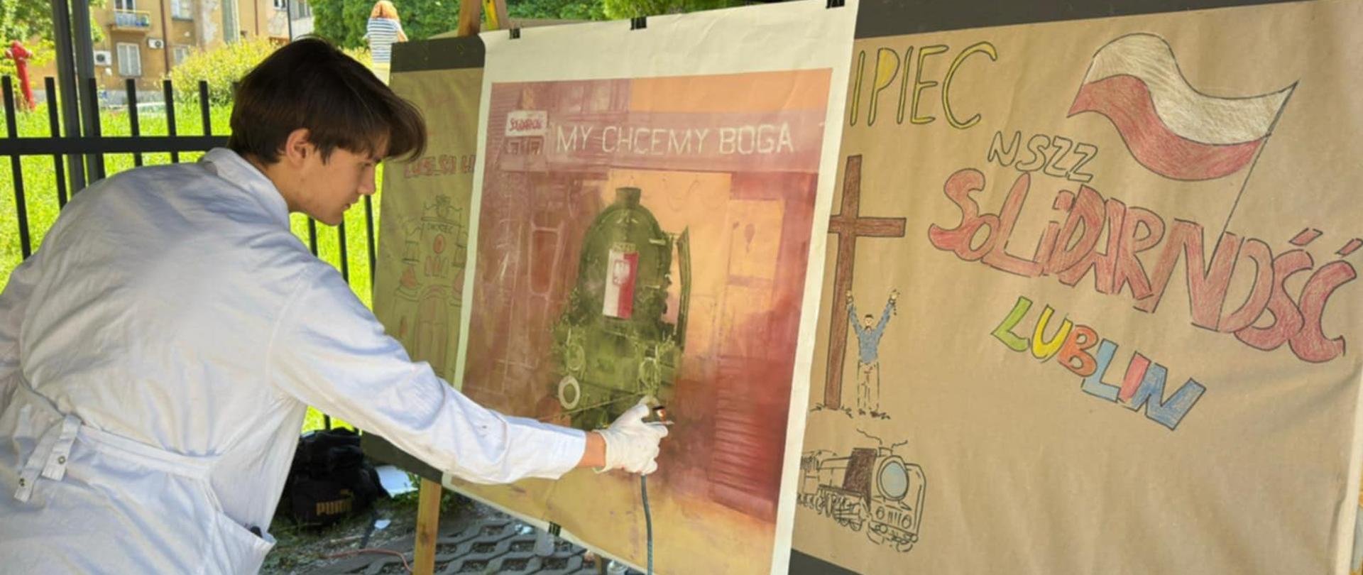 Na zdjęciu widać młodego chłopaka w białym fartuchu malarskim, pracującego nad dużym plakatem przy pomocy aerografu (pistoletu natryskowego). Znajduje się pod namiotem, na zewnątrz, obok metalowego ogrodzenia. Plakat, nad którym pracuje, przedstawia historyczną lokomotywę z napisem „MY CHCEMY BOGA” oraz logiem „Solidarność” i nawiązaniami do stanu wojennego i walki o wolność w Polsce. Obok znajduje się drugi plakat z hasłem „1 PIEC NSZZ Solidarność LUBLIN”, rysunkiem biało-czerwonej flagi, krzyża, człowieka w geście zwycięstwa oraz lokomotywy. Tło zdjęcia pokazuje zieloną trawę, drzewa oraz fragmenty budynków mieszkalnych.