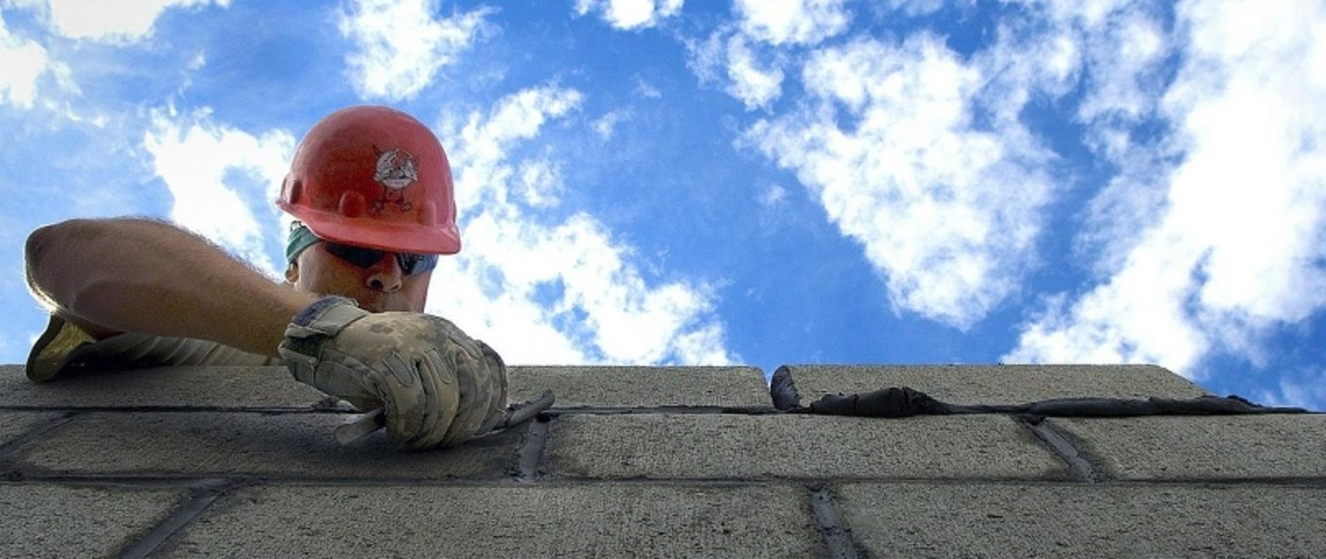 Robotnik w czerwonym kasku układający cegły; w tle niebieskie niebo z chmurami.