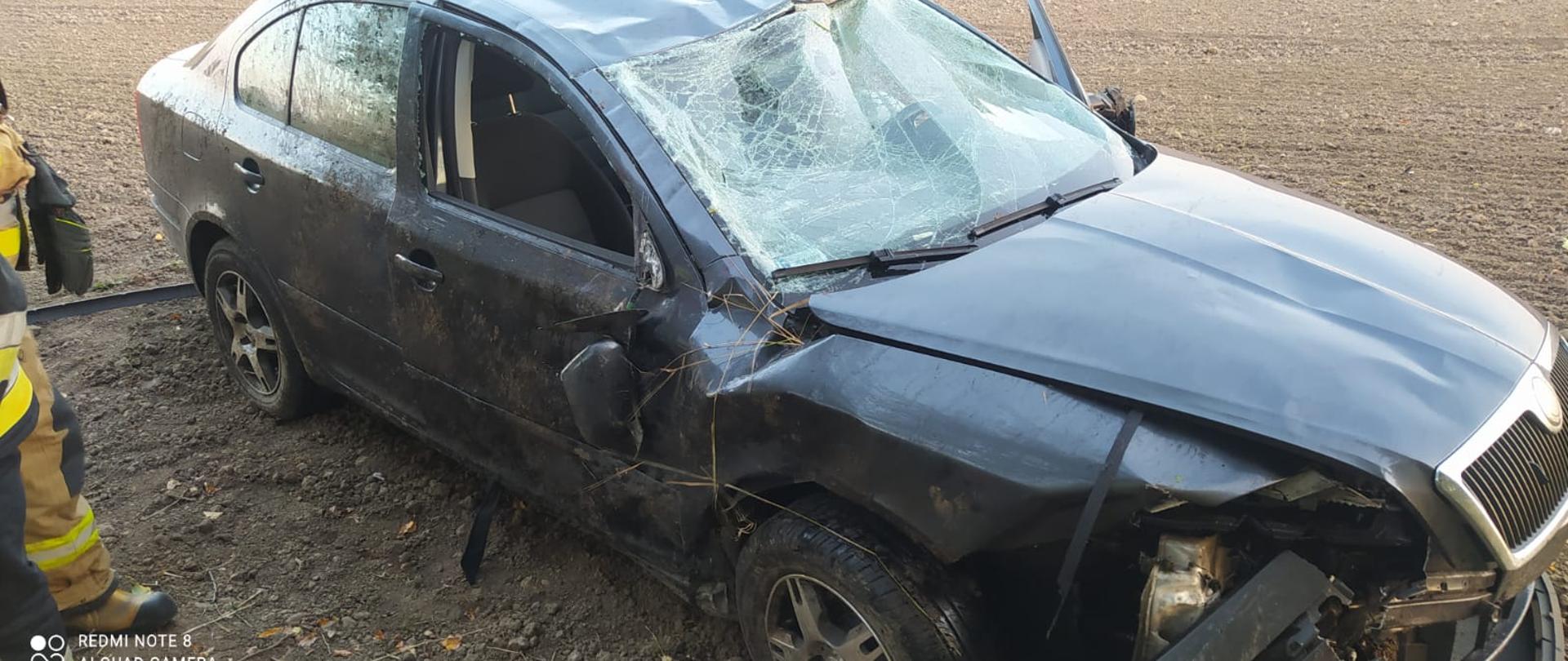 Na polu stoi auto noszące ślady dachowania. Ma rozbitą przednią szybę oraz uszkodzenia po bokach od toczenia się po skarpie.