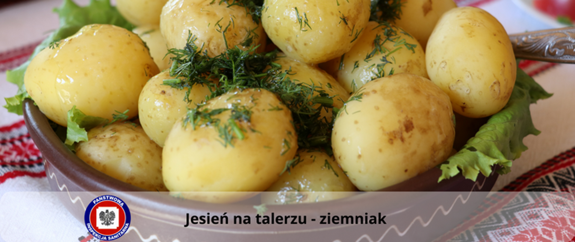 Na kolorowym obrusie stoi brązowa miska z ugotowanymi w całości ziemniakami, polanymi tłuszczem, posypanymi koperkiem i przyozdobionymi liśćmi zielonej sałaty. Na dole jasny przezroczysty pasek z ciemnym napisem Jesień na talerzu - ziemniak, z jego lewej strony logo Państwowej Inspekcji Sanitarnej.