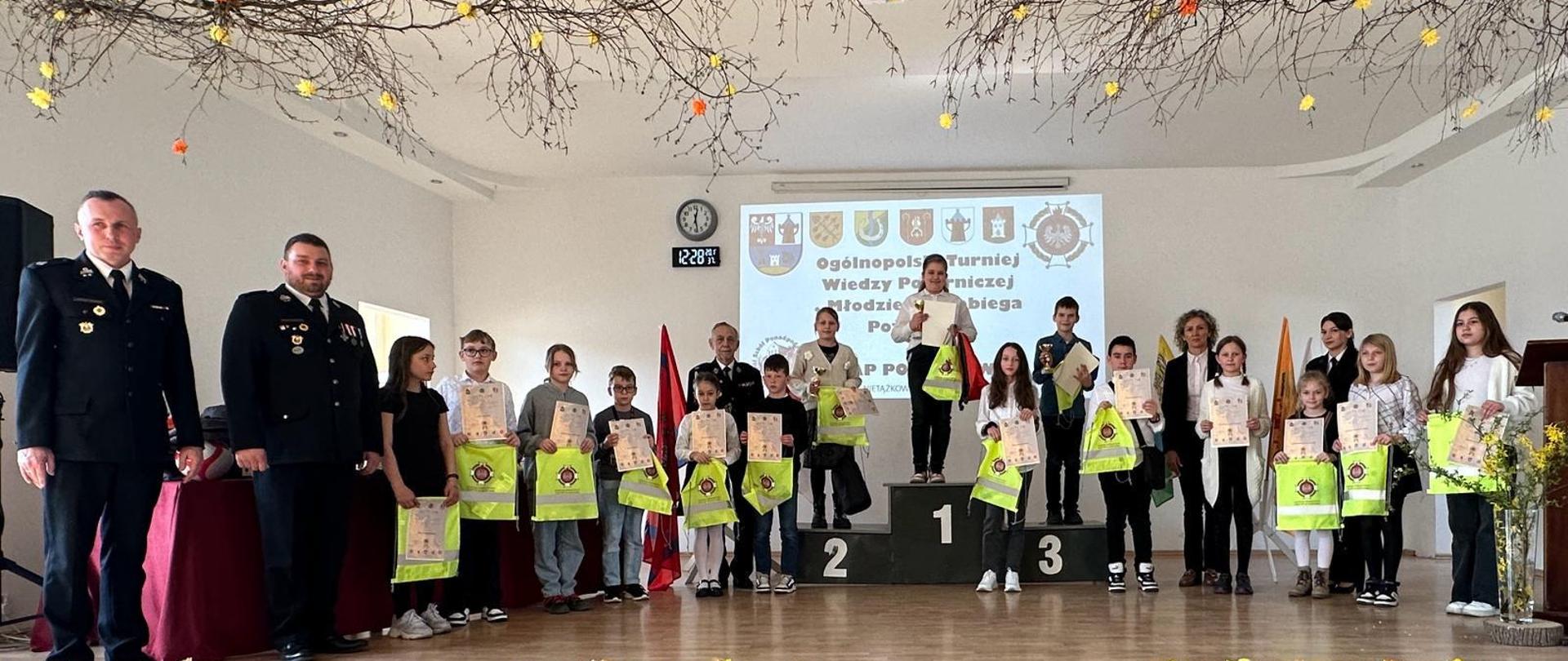Ogólnopolski Turniej Wiedzy Pożarniczej 2024 - etap powiatowy. Młodzież podczas dekoracji zwycięzców, osoby stoją na podium i w rzędzie z dyplomami i upominkami.