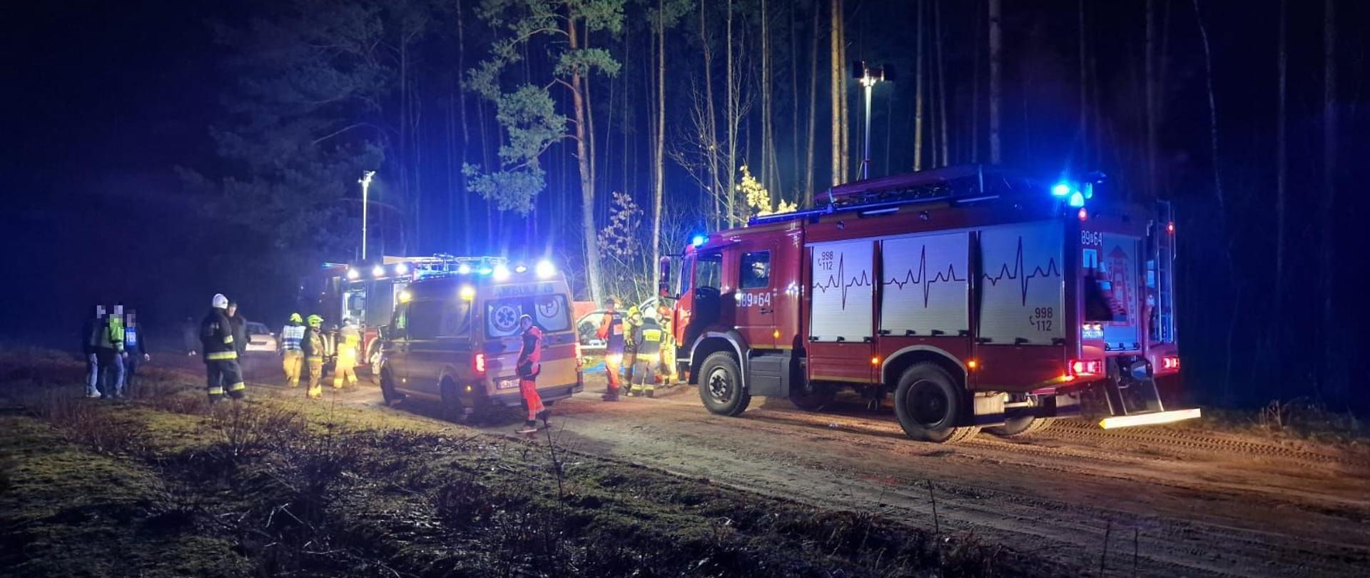 samochody strażackie stoją z podniesionym najaśnicami oświetlając miejsce zdarzenia pomiędzy nimi stoi karetka pogotowia ratunkowego w okół las dookoła samochodów strażacy 
