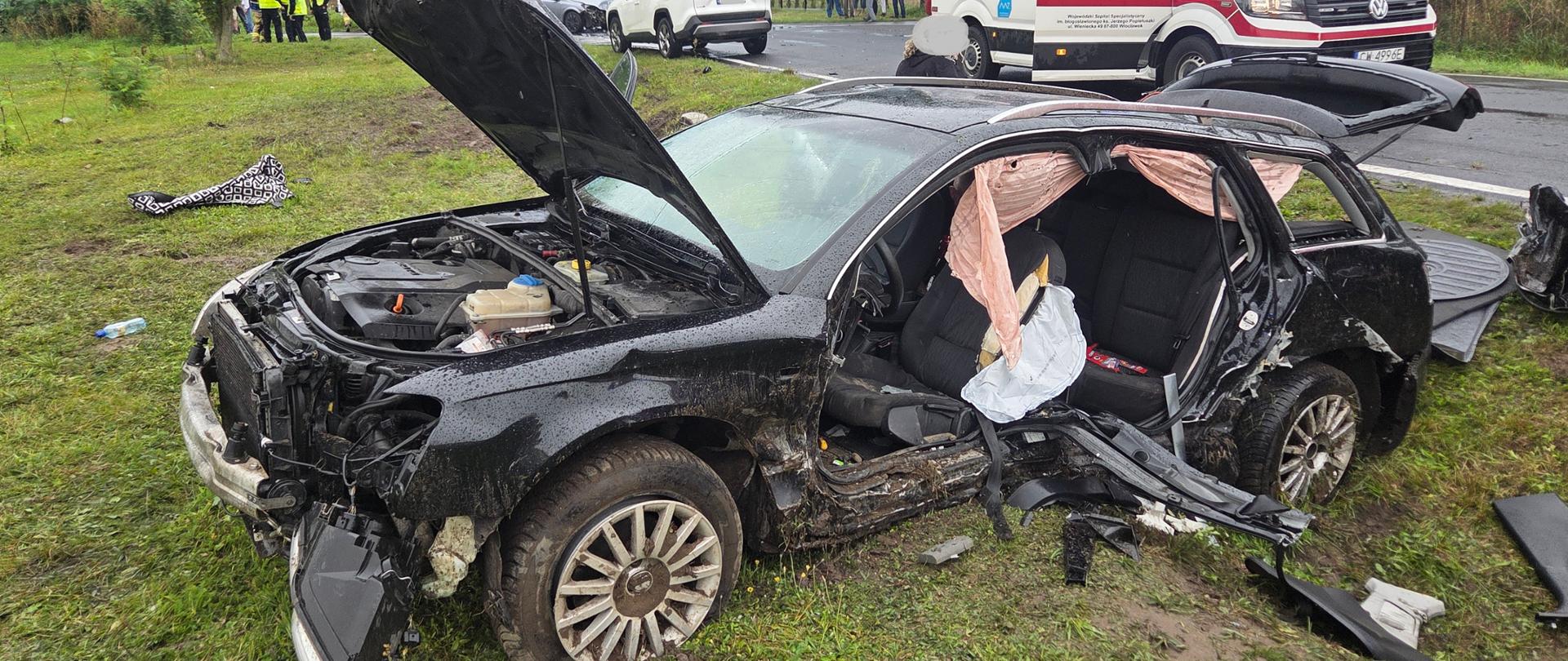 Samochód stoi w przydrożnym rowie. Uszkodzony przód oraz bok samochodu. Poduszki wystrzeliły. Na jezdni stoi samochód strażacki oraz karetka pogotowia. 