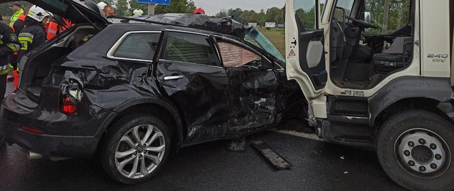 Zdjęcie przedstawia widoczny od strony pasażera uderzony w bok czarny pojazd marki Mazda przez biały samochód dostawczy marki Volvo