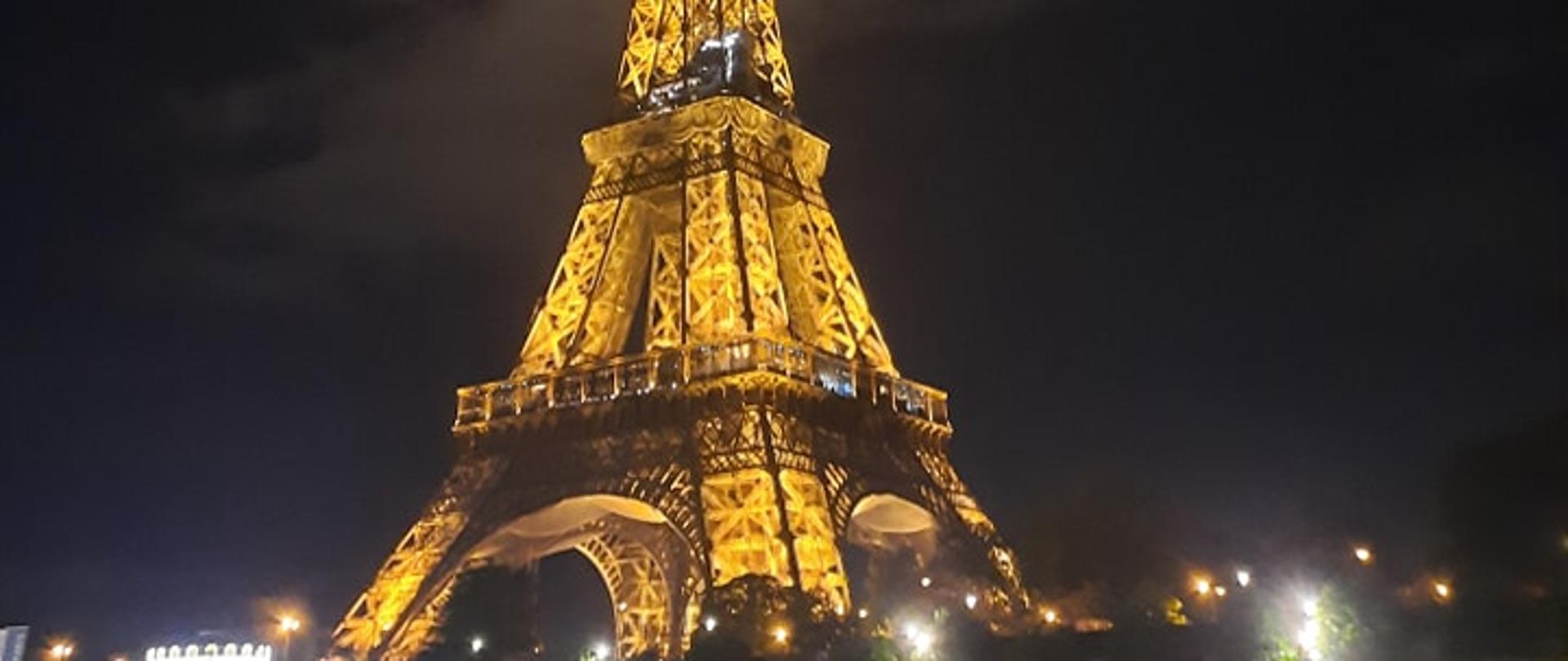 Zdjęcie wieży Eiffla nocą. Przed wieżą z lewej i prawej strony na rzece 2 statki wycieczkowe. W tle za wieżą widoczne chmury.