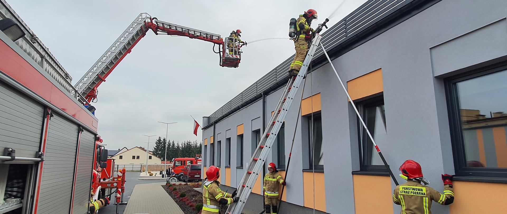 strażacy JRG Kozienice podczas ćwiczeń, jeden strażak w koszu podnośnika podaje prąd wody na dach budynku, drugi strażak stoi na drabinie i podaje prąd wody na dach budynku, trzech strażaków asekuruje drabinę, na której stoi strażak, 