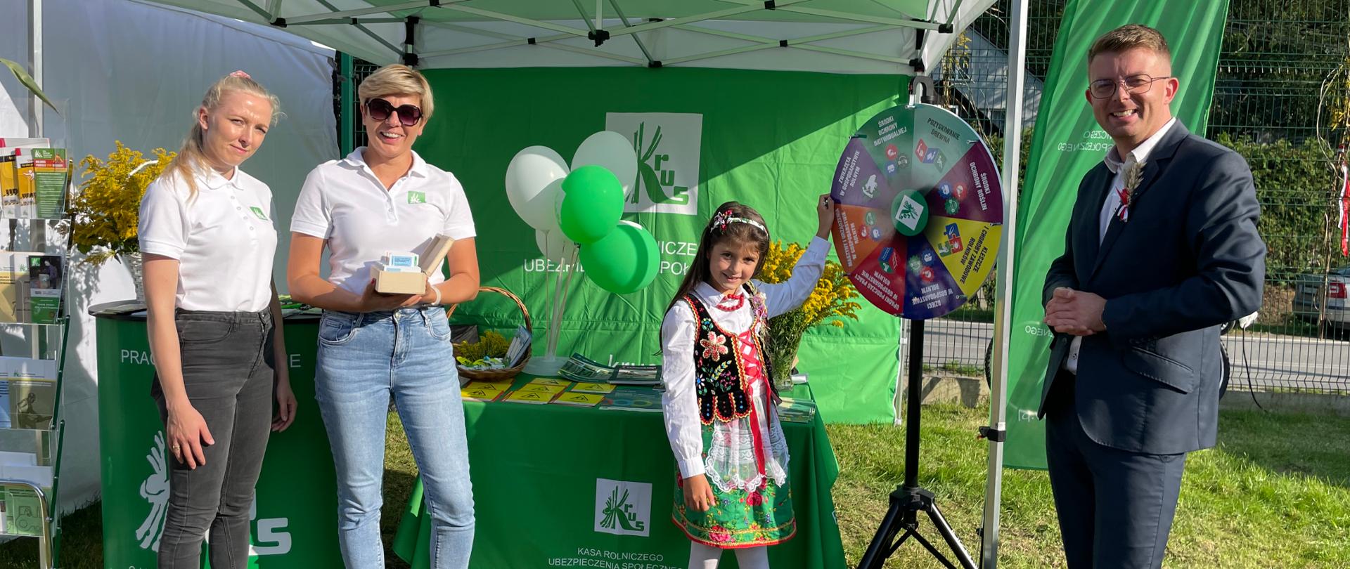 Na tle stoiska informacyjnego stoi mężczyzna, dwie kobiety i dziewczynka w stroju ludowym, która kręci Kołem Fortuny