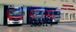 Przed budynkiem remizy strażacy stoją na zbiórce. Za nimi w bramach garażowych widoczne samochody z włączonymi sygnałami świetlnymi i dźwiękowymi. 