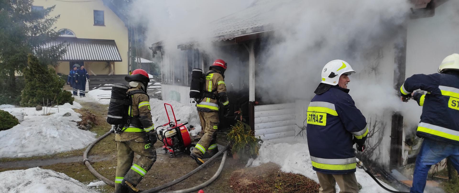 Na tle białego budynku po lewej stronie dwóch strażaków w aparatach OUO wprowadza linię gaśniczą po prawej stronie dwóch strażaków prowadzi prace rozbiórkowe.