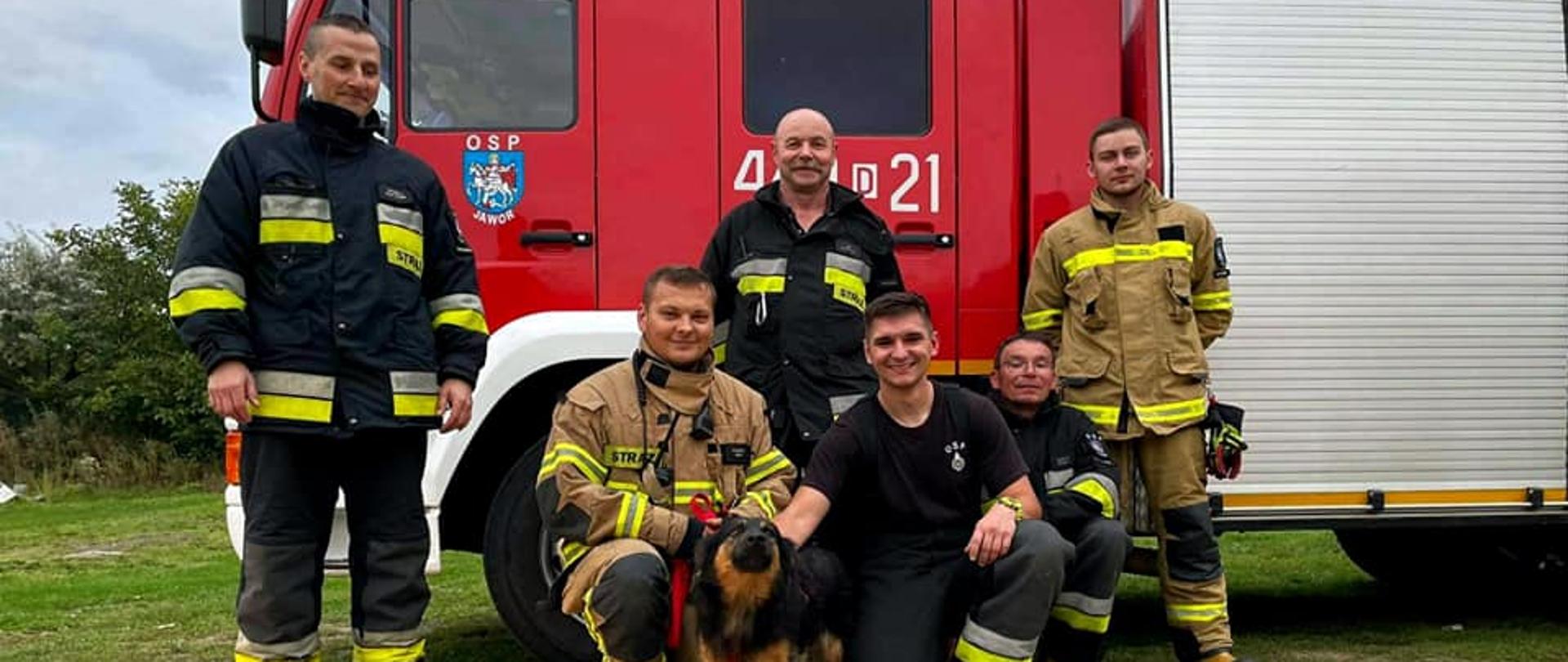 Obraz przedstawia strażaków z uratowanym psem na tle samochodu pożarniczego.