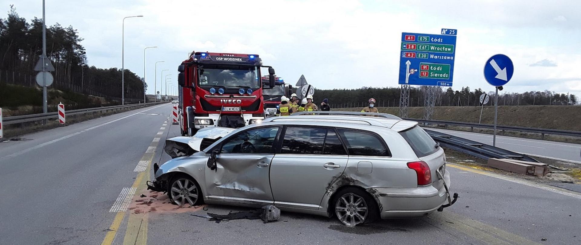 zdjęcie przedstawia uszkodzony podczas kolizji samochód osobowy koloru srebrnego, przy przednim prawym kole rozsypany jest sorbent, za ww. pojazdem stoją samochody ratowniczo gaśnicze straży pożarnej , oraz ratownicy 