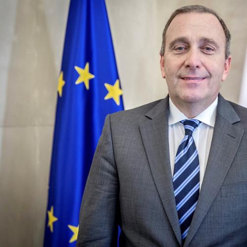 Grzegorz Schetyna kolorowy portret mężczyzny stojącego na tle biało-czerwonej polskiej flagi po prawej i flagi unijnej niebieskiej w żółte gwiazdy po lewej, siwiejące włosy, szara marynarka, granatowy krawat w niebieskie paski, biała koszula.