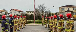 Strażacy stoją w dwóch rzędach na przeciwko siebie, uroczyście podnosząc flagę Rzeczypospolitej Polskiej na maszt.