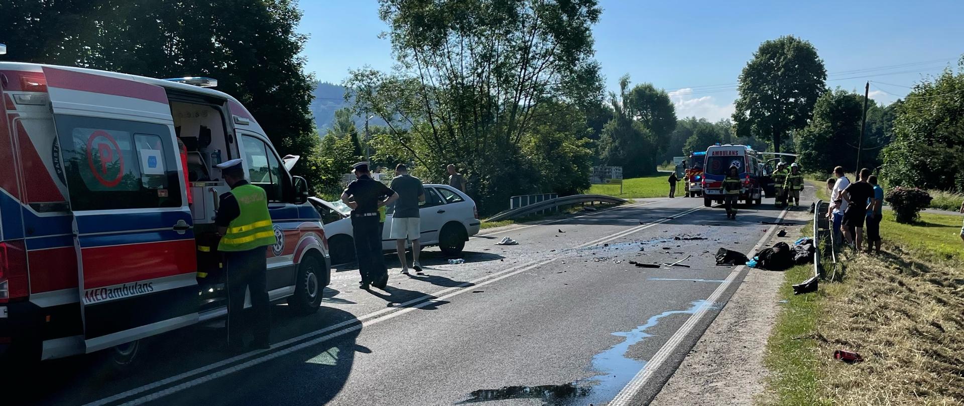 Na pierwszym planie karetka pogotowia ratunkowego, obok niej stoi umundurowany policjant. Na dalszym planie po lewej stronie uszkodzony pojazd, który brał udział w zdarzenia. Na jezdni leżą porozbijane elementy pojazdów. Po prawej stronie stoją postronne osoby, które obserwują pracę strażaków policji i ratowników medycznych. Na najdalszym planie widoczna druga karetka pogotowia ratunkowego, samochód straży pożarnej oraz śmigłowiec LPR. 