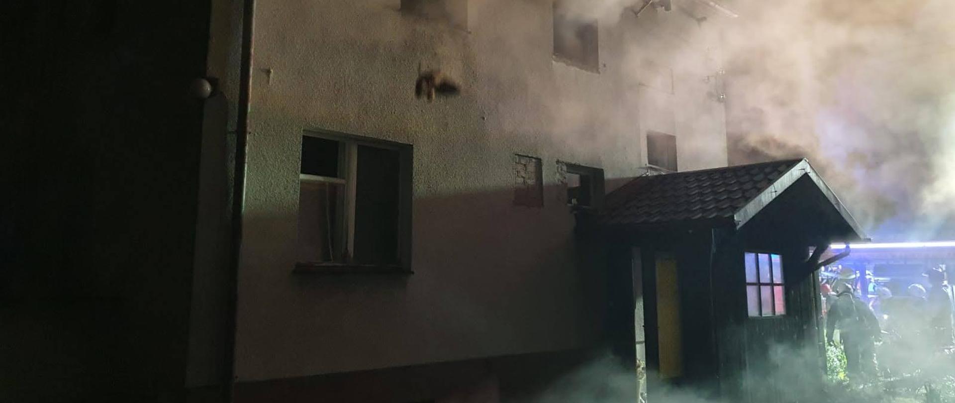 Pora nocna, widoczne kontury budynku mieszkalnego murowanego, z widocznym zadaszeniem nad wejściem. Budynek otoczony kłębami dymu. 