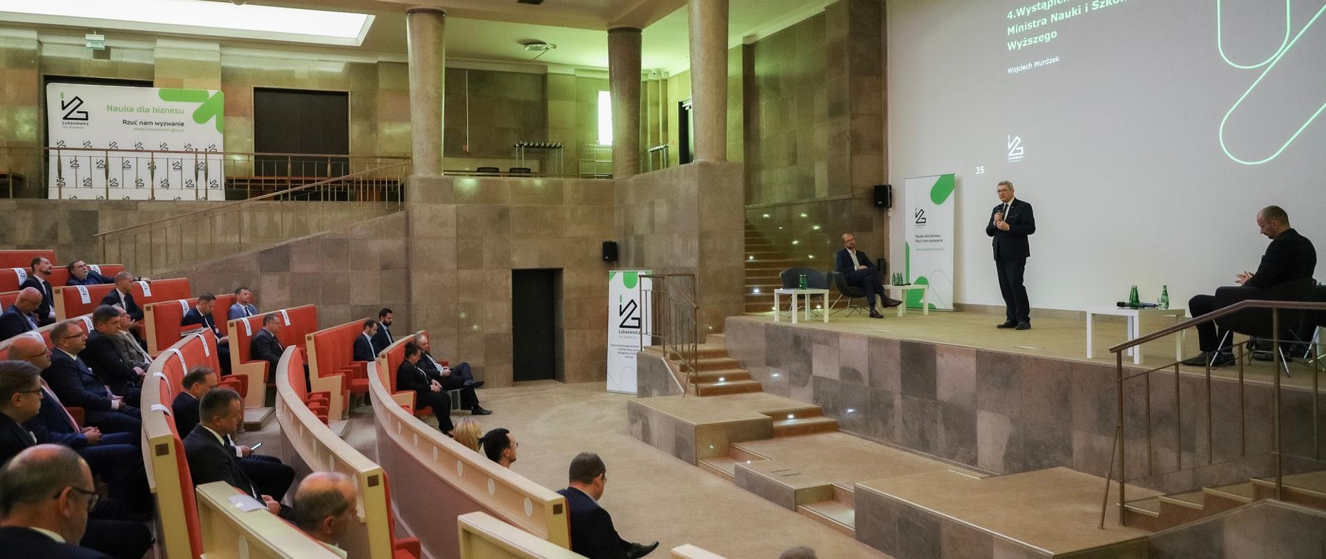 W dużej sali minister przemawia do mikrofonu, przed nim siedzą dyrektorzy.