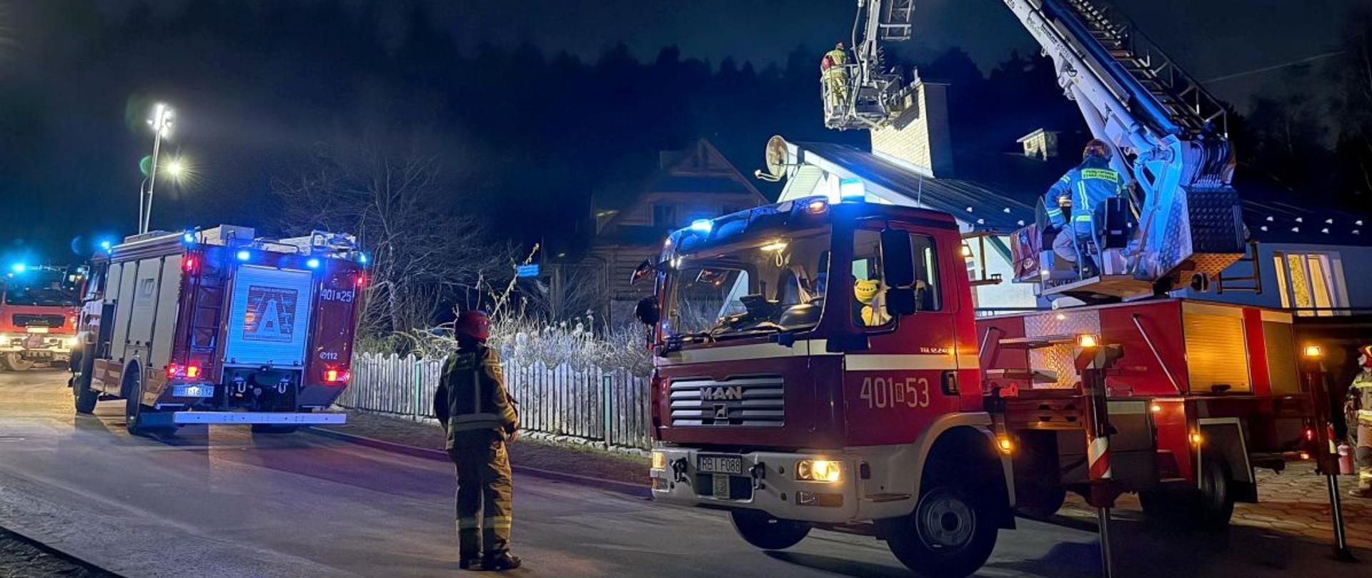 Na zdjęciu widać trzy samochody strażackie. Dwa z nich to samochody gaśnicze a trzeci to drabina z koszem która w tym przypadku jest rozłożona a w koszu znajduje się strażak nad kominem domu jednorodzinnego. Przy samochodzie z drabiną stoi strażak w ubraniu specjalnym. 