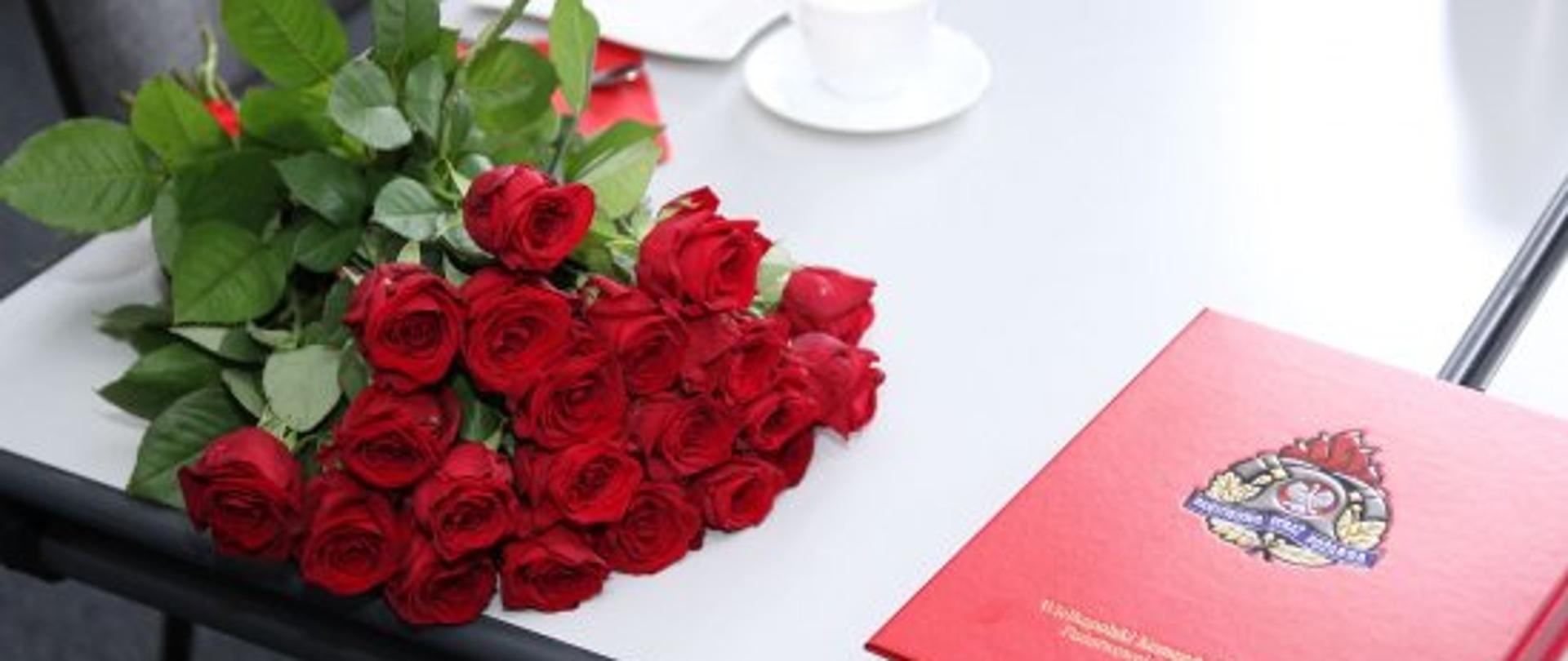 na zdjęciu widoczny jest duży bukiet z czerwonych róż leżący na szarym stole obok czerwonej teczki z logo państwowej straży pożarnej oraz białej filiżanki do kawy