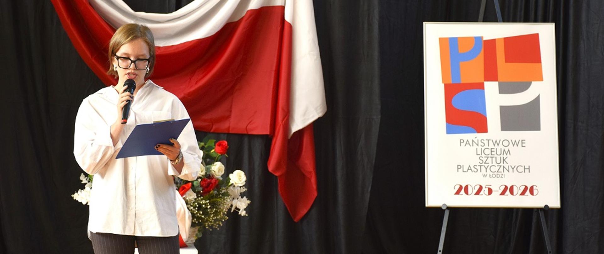 Na zdjęciu przewodnicząca szkoły wita społeczność szkolną. W tle zawieszona flaga Polski. Na sztaludze logotyp szkoły