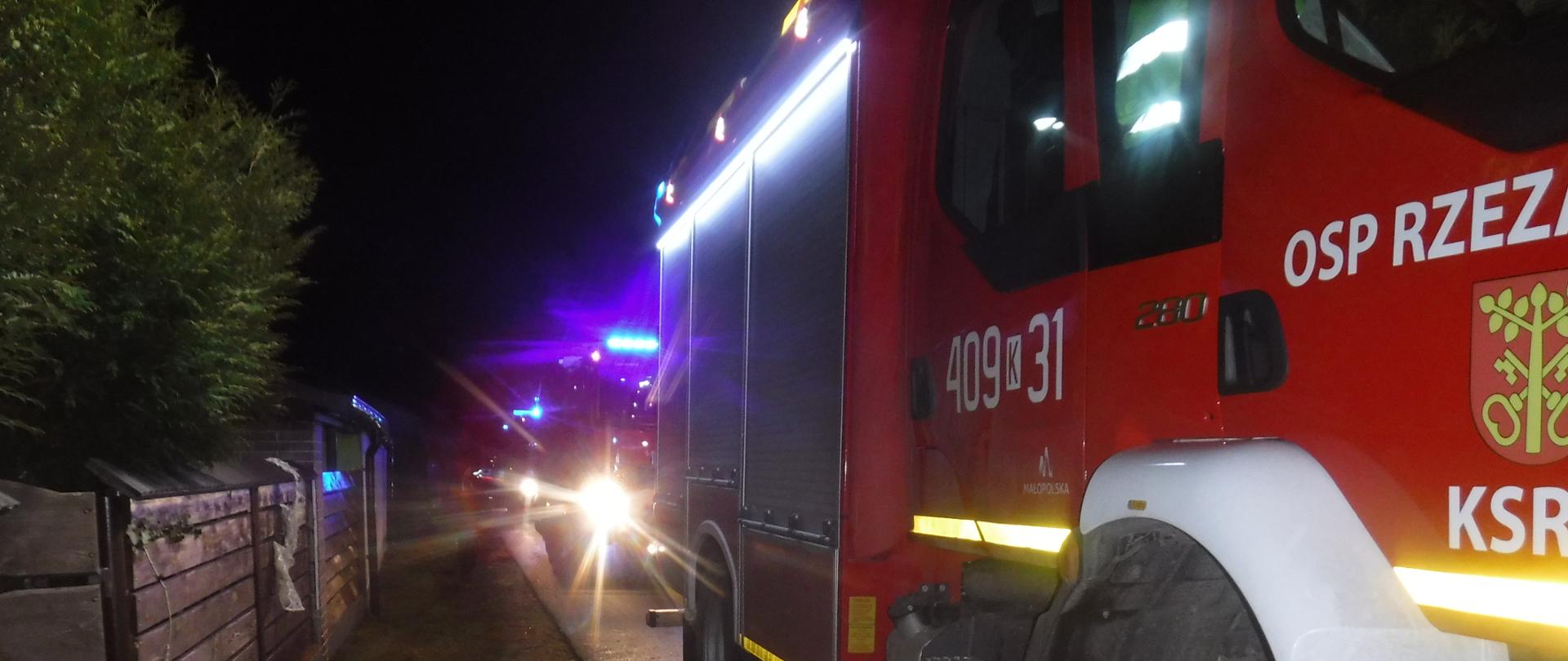 Zdjęcie przedstawia stojące trzy czerwone samochody pożarnicze ustawione jeden za drugim. Jest pora Nocna. Pierwszy samochód na drzwiach ma numer operacyjny 409K31 oraz napis OSP Rzezawa herb i napis KSRG. Wszystkie auta mają włączone światła drogowe oraz niebieskie światła błyskowe. Po lewej stronie widać drewniane ogrodzenie w kolorze brązowym.