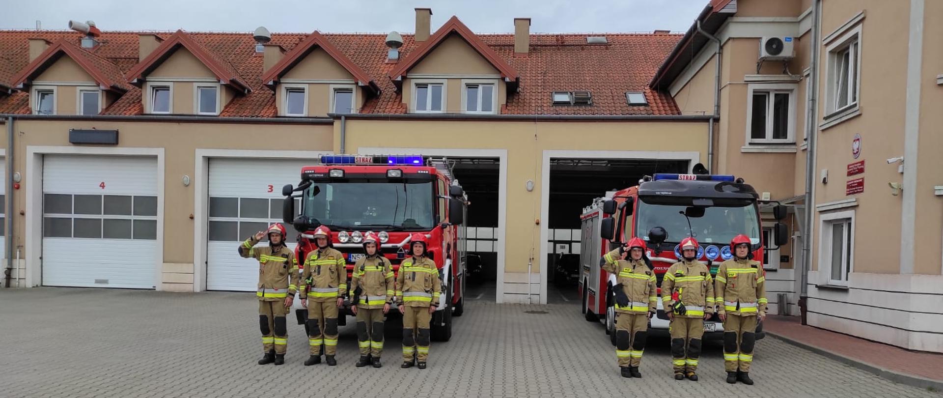 Zdjęcie przedstawia strażaków oddających honor z okazji rocznicy Powstania Warszawskiego na tle samochodów pożarniczych oraz budynku Komendy Powiatowej PSP w Szczytnie