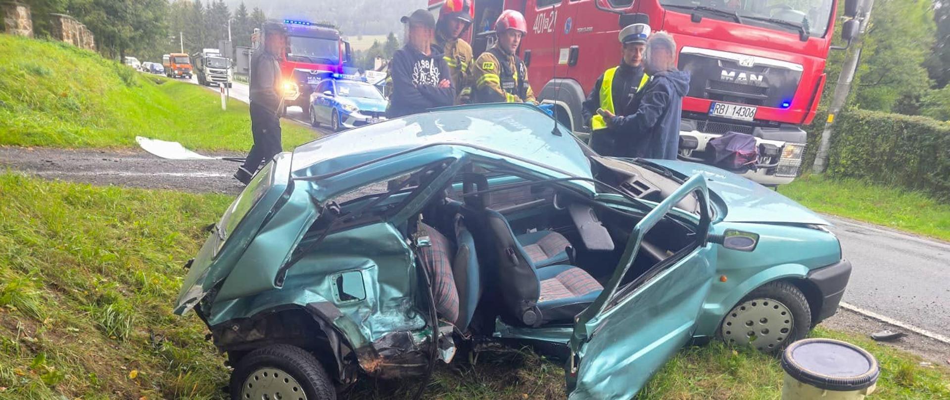 Zdjęcie przedstawia samochód po wypadku z wgniecionym prawym bokiem poza drogą w rowie. Za samochodem znajdują się samochody strażackie i radiowóz oraz strażacy, policjant i osoby postronne. 