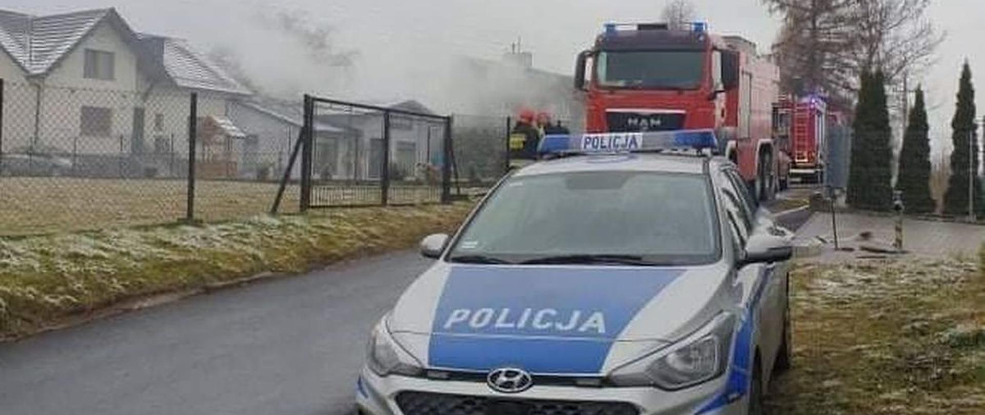 Widok z przodu. Na pierwszym planie radiowóz Policji. Na drugim planie samochody ratowniczo-gaśnicze, strażacy Państwowej Straży Poarnej oraz zadymione zabudowania.