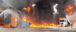Pożar hali murowanej oraz samochodu ciężarowego z naczepą znajdującego się w pobliżu. Strażak podaje jeden prąd piany na palący się samochód.