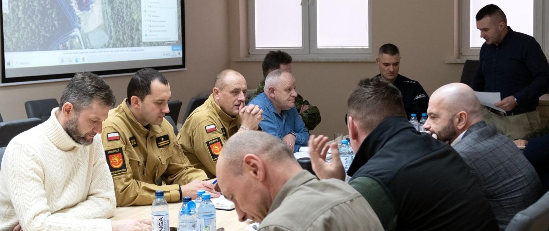 Zdjęcie wykonane w budynku starostwa bieszczadzkiego. Znajdują się na nim przedstawiciele służb: straż pożarna, policja, wojsko oraz pracownicy starostwa i przedstawiciele władz samorządowych. Straż pożarną reprezentują komendant powiatowy oraz zastępca komendanta powiatowego. 