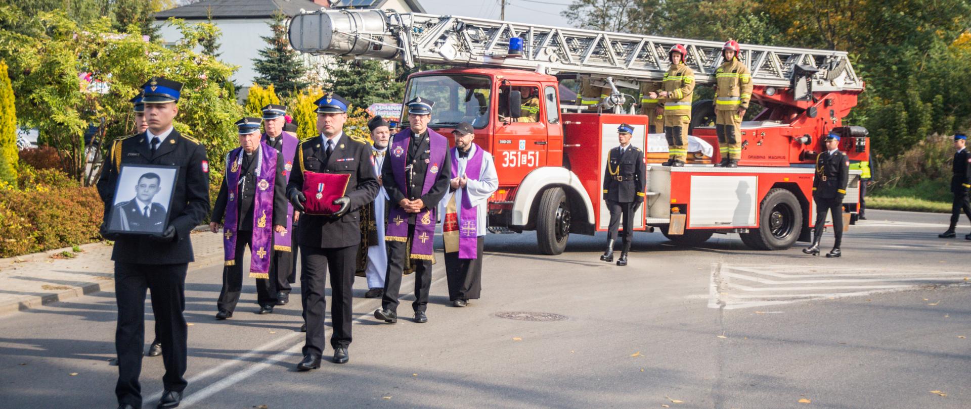 Strażacy niosą fotografię zmarłego funkcjonariusza i nadane Mu medale, za nim w kondukcie idzie ksiądz oraz jedzie samochód z drabiną pożarniczą