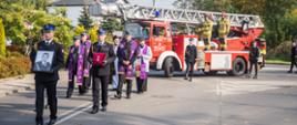 Strażacy niosą fotografię zmarłego funkcjonariusza i nadane Mu medale, za nim w kondukcie idzie ksiądz oraz jedzie samochód z drabiną pożarniczą