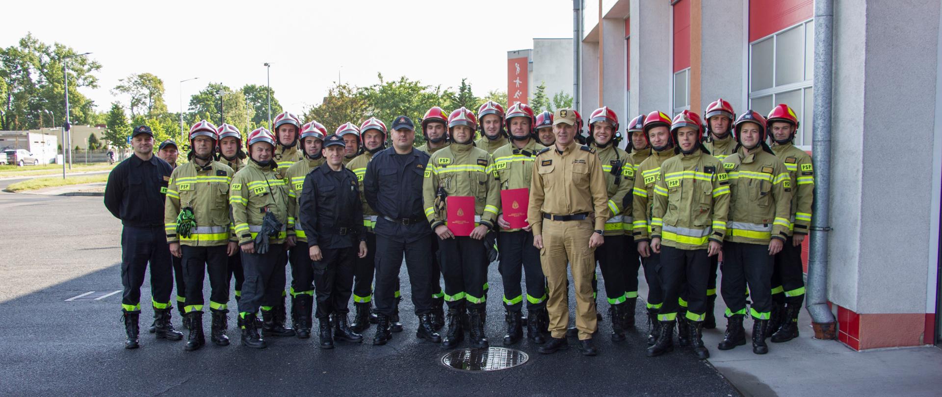 Strażacy w mundurach stoją w szeregu obok budynku komendy.