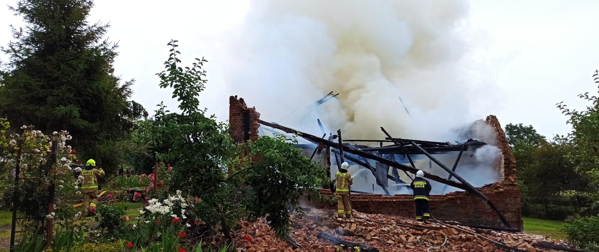 Dwóch strażaków ochotników podaje prądy wody na zniszczony dach palącego się budynku gospodarczego. Nad budynkiem unoszą się kłęby dymu.