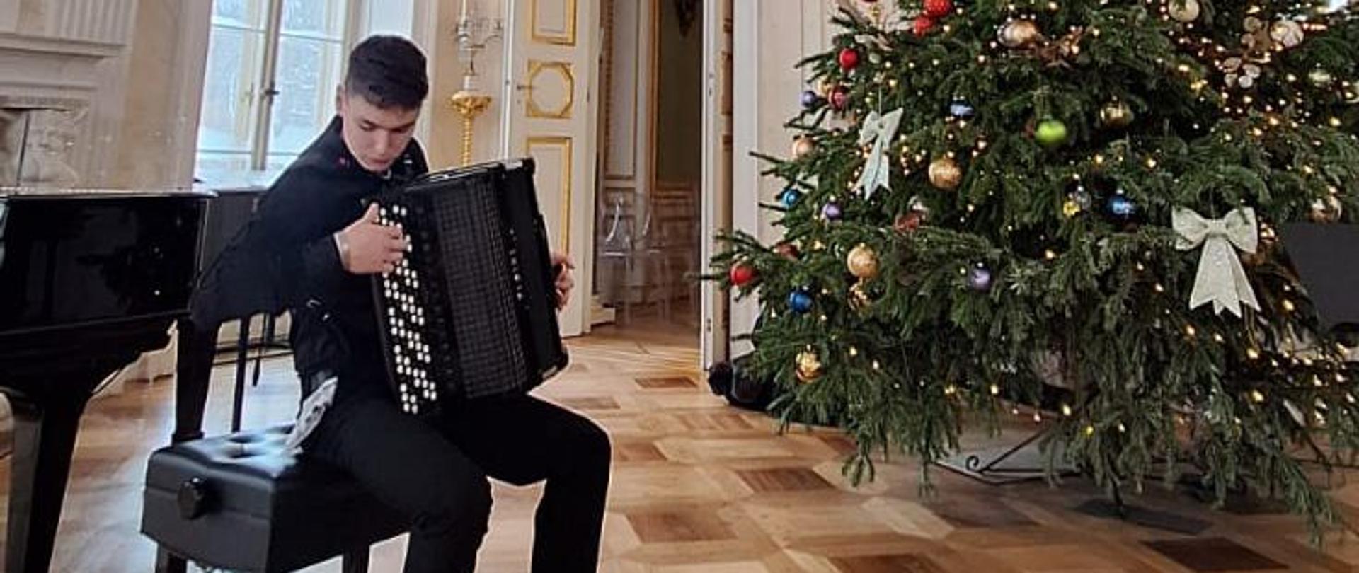 Zdjęcie - Mateusz Neckar na próbie przed recitalem w Pałacu Łazienkowskim. Mateusz gra na akordeonie w sali pałacowej, w tle choinka.