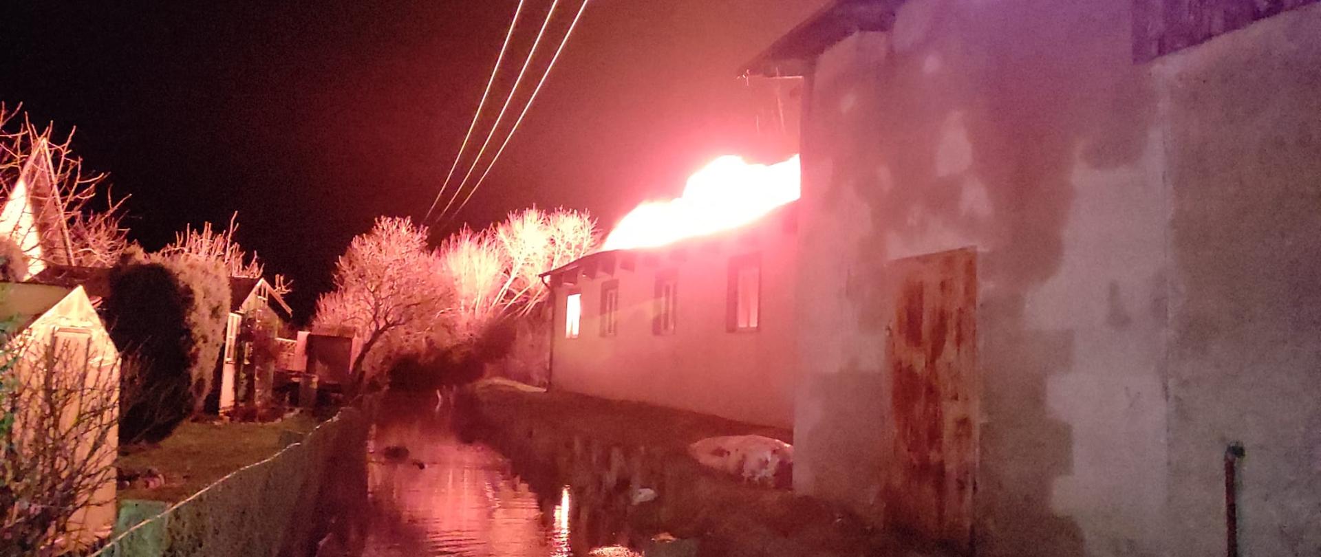 Noc. Z prawej strony budynek. dach w ogniu. na środku rowem płynie woda.