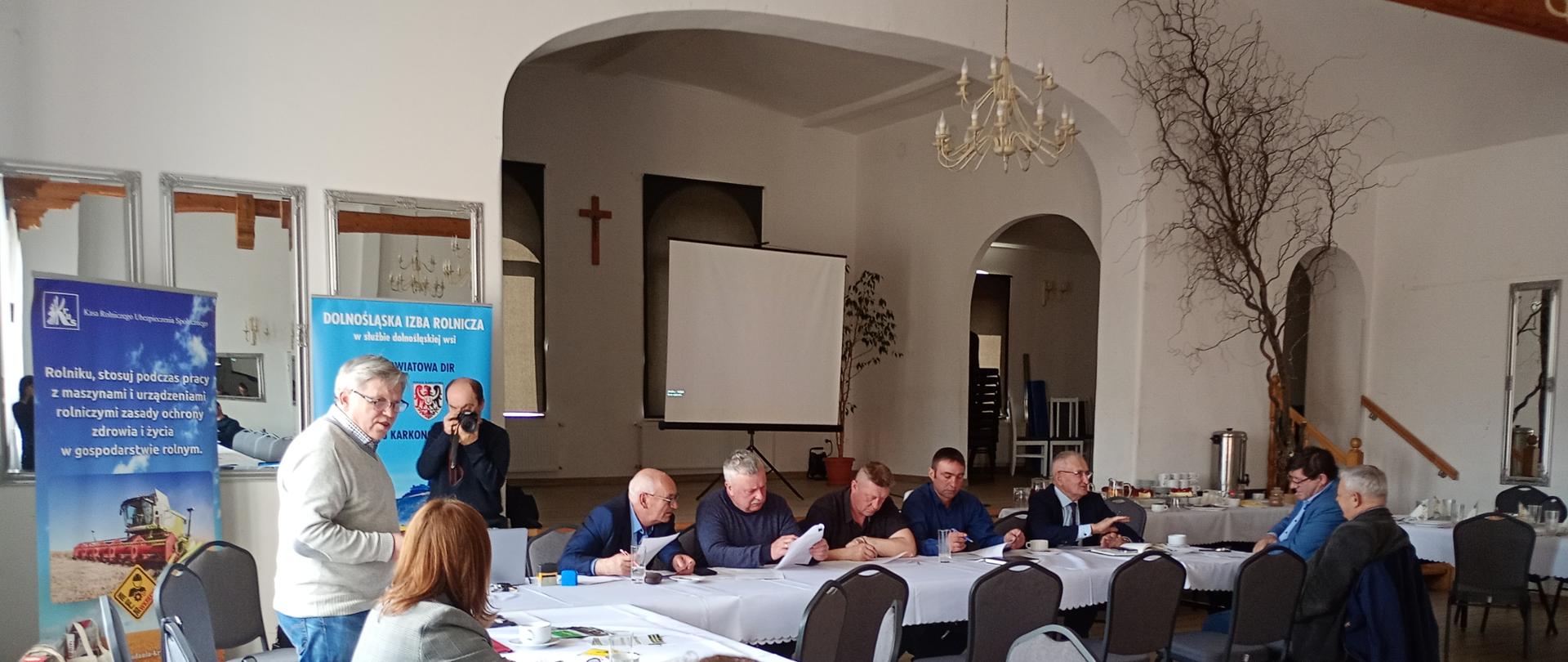 Uczestnicy posiedzenia siedzący przy dużym stole w kształcie litery "L". Kierownik Oddziału w Jeleniej Górze stoi i przemawia do uczestników posiedzenia.