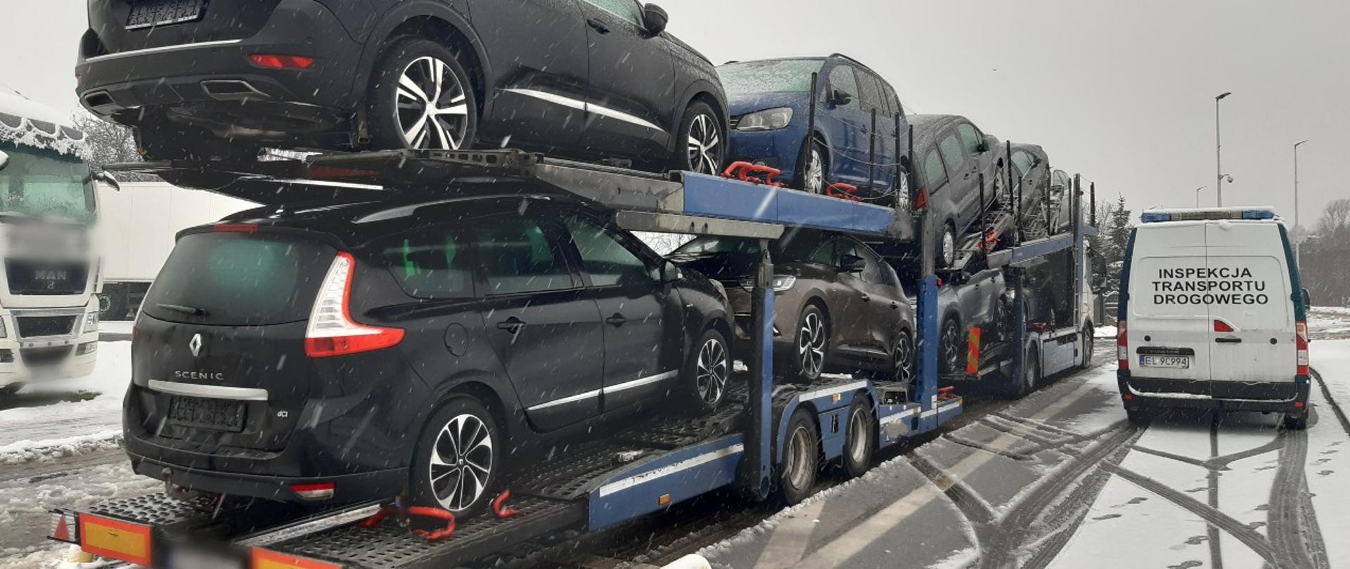 Laweta załadowana kilkoma samochodami osobowymi stoi na ośnieżonym parkingu. Obok niej znajduje się biały bus Inspekcji Transportu Drogowego z włączonymi światłami ostrzegawczymi. Na ziemi widać ślady opon oraz padający śnieg.
