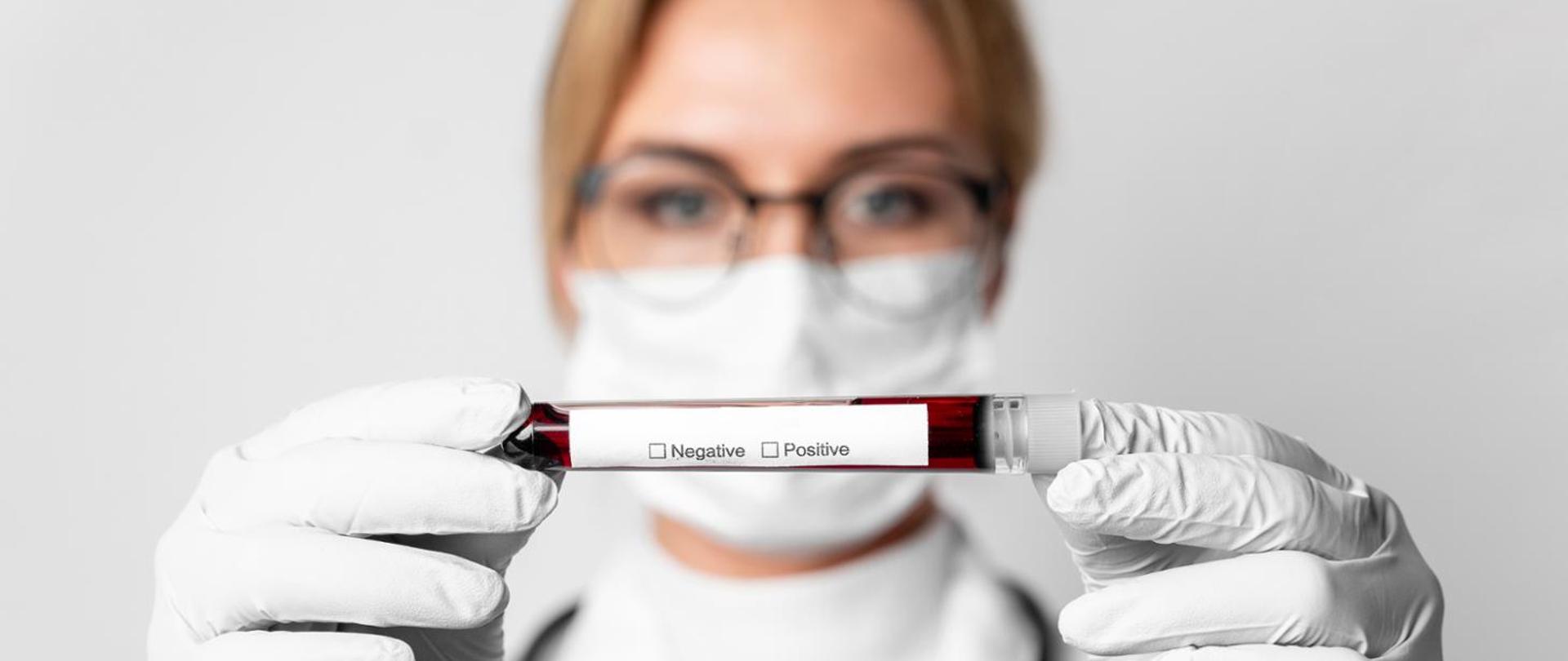 kobieta w maseczce i odzieży ochronnej trzymająca w ręku fiolkę z pobraną krwią na test na HIV