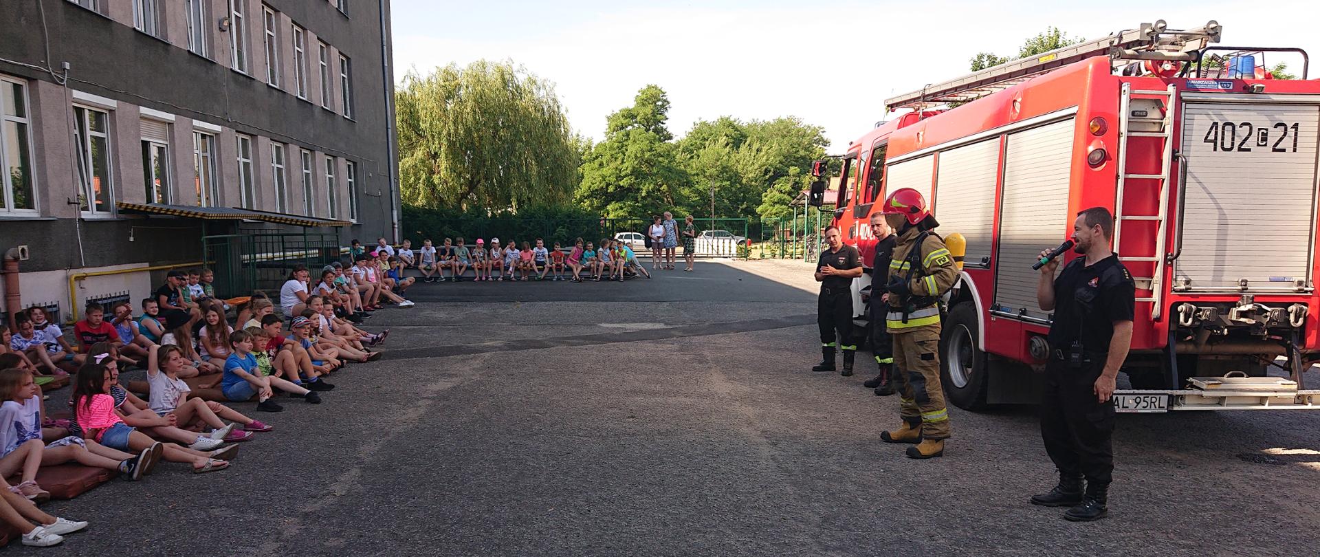 Na zdjęciu po prawej stronie widoczny jest wóz strażacki z JRG Ciechocinek, przed którym stoją strażacy prowadzący prelekcję na temat bezpiecznych wakacji. Trzech strażaków ubranych jest w ubrania koszarowe, podczas gdy jeden jest ubrany w pełne ubranie specjalne, wraz z aparatem OUO, w celu zaprezentowania dzieciom używanego przez strażaków sprzętu. Przy budynku szkoły z lewej stronie zdjęcia widać siedzących na materacach i ławkach uczniów szkoły podstawowej.