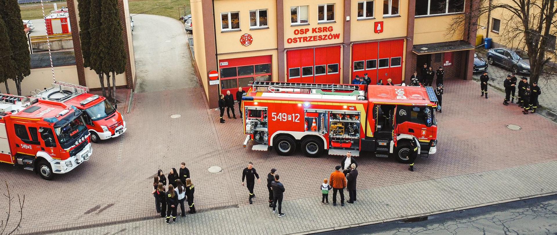 Zdjęcie przestawia samochody OSP Ostrzeszów, w tym po prawej stronie stoi nowy pojazd. Dookoła stoją ludzie zainteresowani wydarzeniem. Na dalszym planie budynku straży pożarnej.