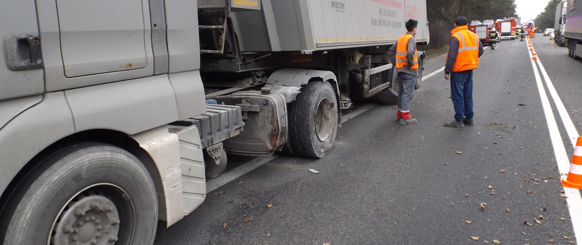 Na zdjęciu znajduje się samochód ciężarowy z lekkimi uszkodzeniami
koła oraz nadkola. W tle widać samochody strażackie, oznakowaną
pachołkami jezdnię, oraz kierowcę samochodu ciężarowego w odblaskowej
kamizelce