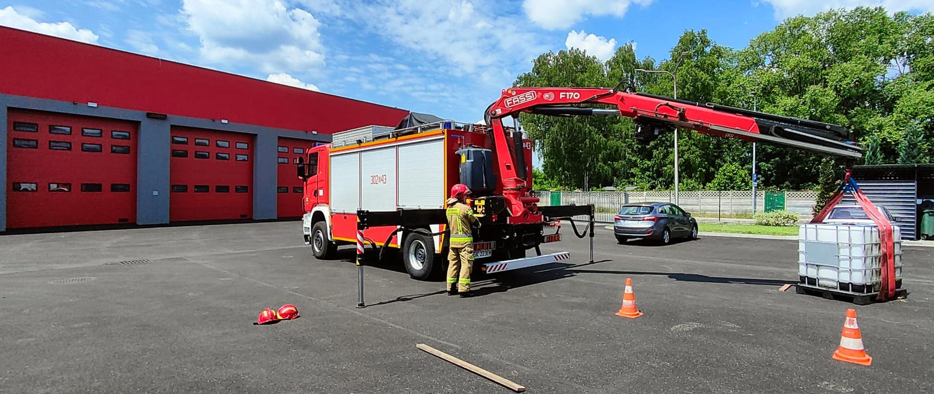 Zdjęcie przedstawia parking na terenie komendy Państwowej Straży Pożarnej. Na parkingu stoi samochód specjalny ratownictwa technicznego z wysuniętym żurawiem, na którym zawieszona jest paleta ze zbiornikiem. Obok samochodu stoi strażak w żółtym ubraniu specjalnym Nomex i chełmie. W tle widoczne czerwone bramy i garaże jednostki.