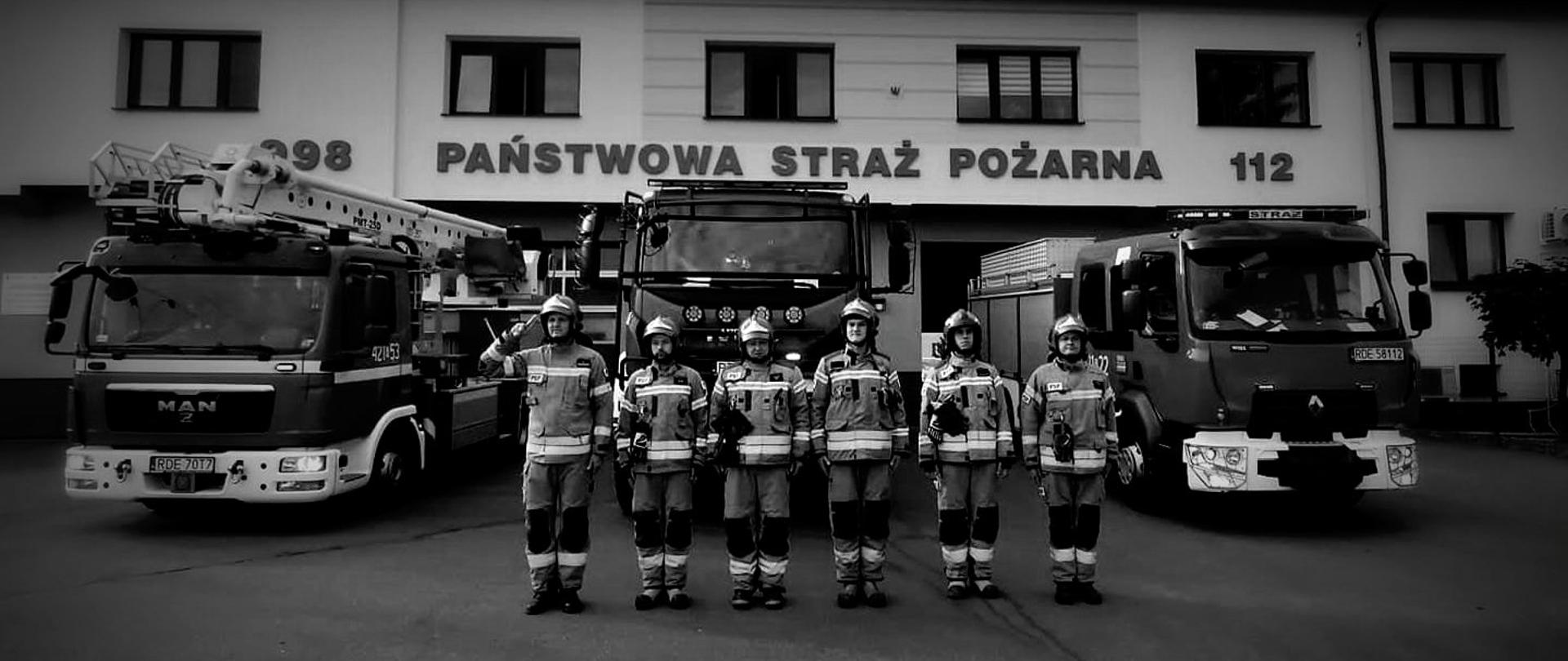 Na zdjęciu sześciu strażakach w ubraniach specjalnych stoi w szeregu. Pierwszy z lewej oddaje salut. Za nimi ustawione samochody pożarnicze. Nad samochodami, na ścianie budynku napis Państwowa Straż Pożarna 112.