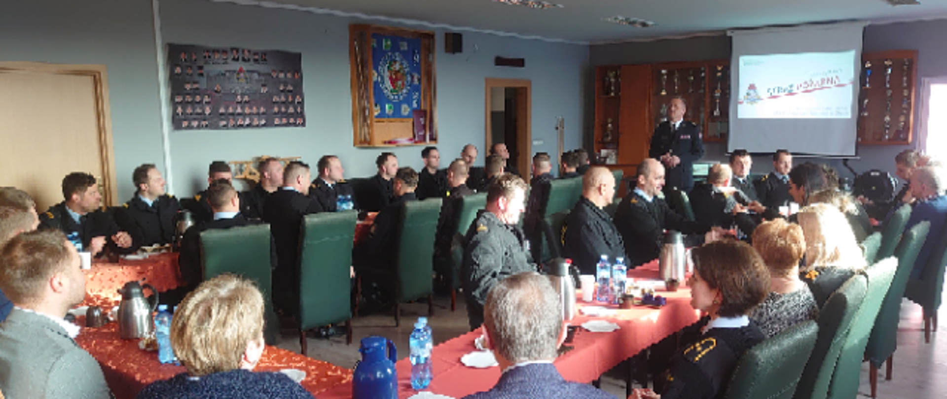 Na zdjęciu znajdują się osoby cywilne oraz funkcjonariusze siedzący za stołem oraz komendant prezentujący osiągnięcia komendy