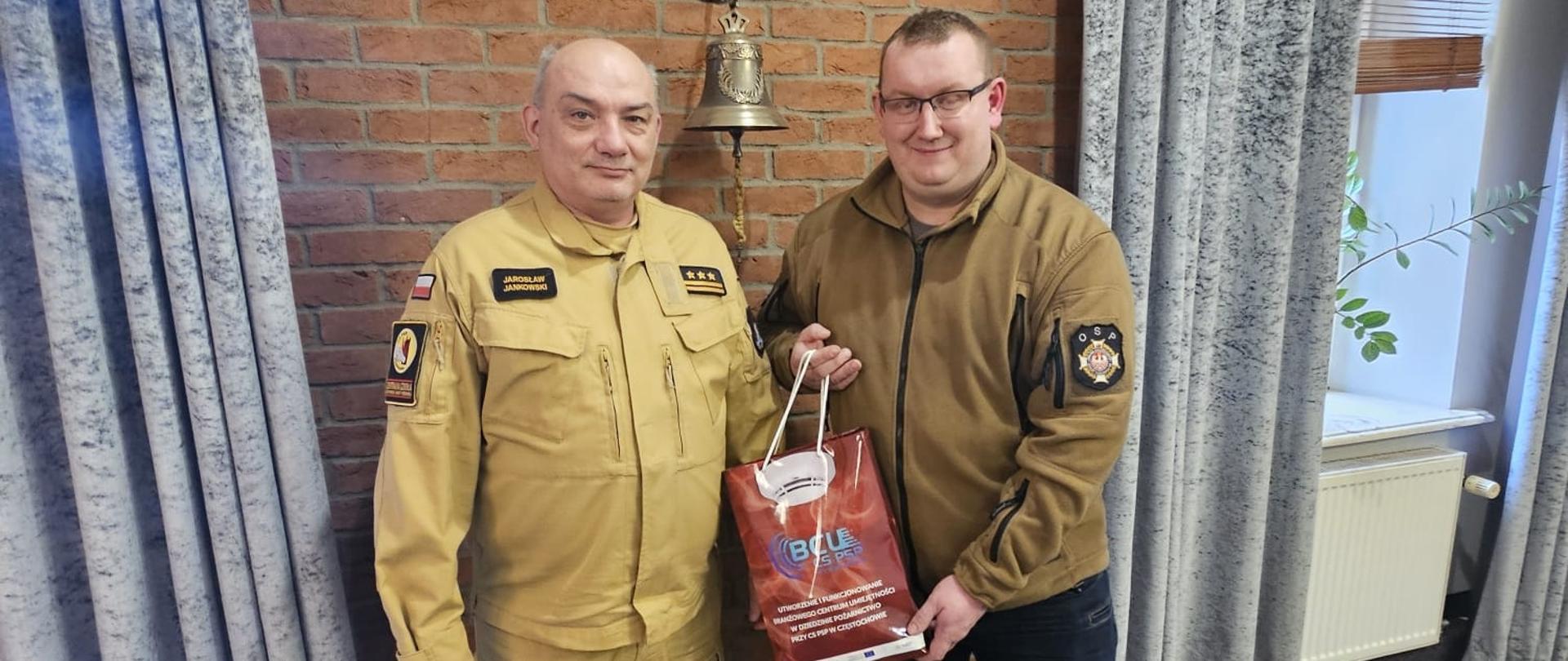 Gabinet Komendanta Centralnej Szkoły PSP, Komendant wraz z gościem pozuje do wspólnego pamiątkowego zdjęcia na tle ceglanej ściany na której wisi metalowe godło oraz dzwonek