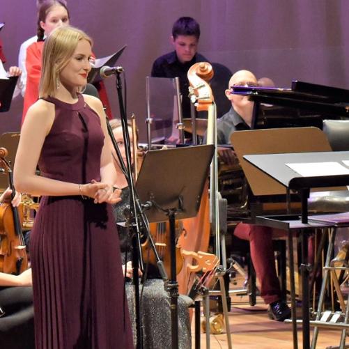 Zdjęcie kolorowe. Wokalistka w amarantowym stroju wieczorowym stoi przy mikrofonie. W tle pulpity, instrumenty i uczniowie orkiestry szkolnej.