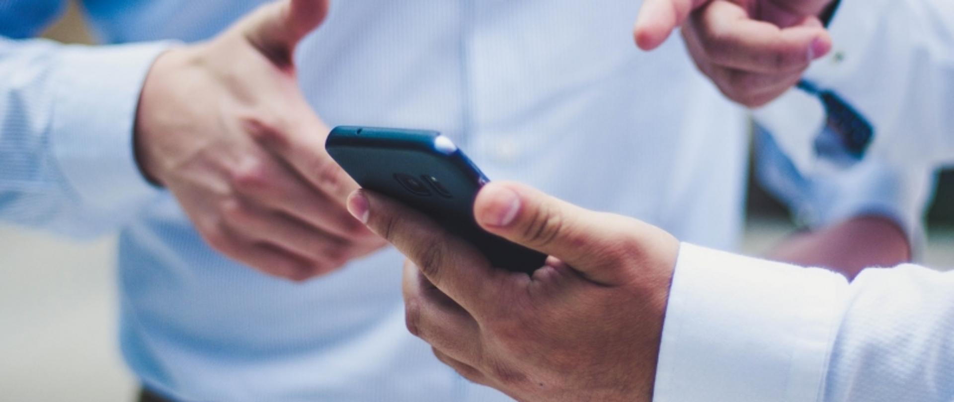 „Trzy osoby w białych koszulach, jedna trzyma smartfon, druga wskazuje na ekran, trzecia pokazuje gest kciuka w górę.”